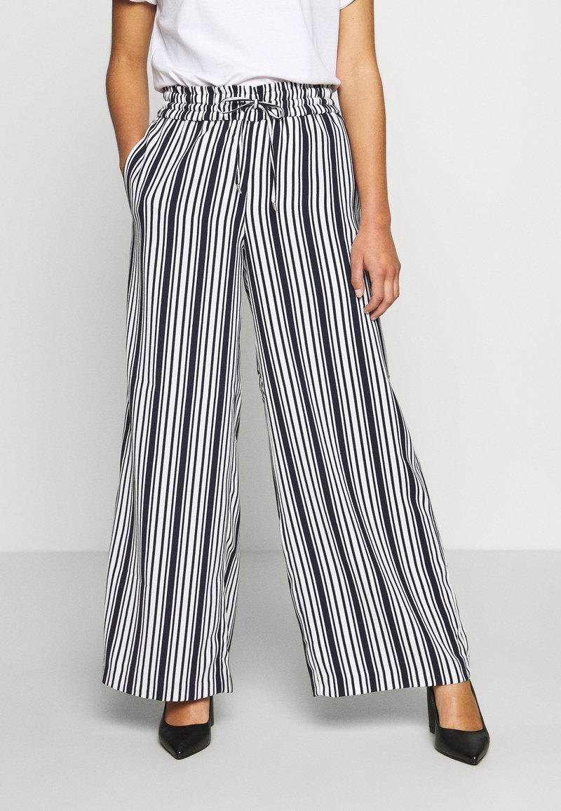 Femme portant un pantalon large à rayures verticales noires et blanches avec une taille à cordon et des talons noirs à bouts pointus.