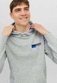 Šedá mikina s kapucí, šňůrkou, přední kapsou a modrými logy na levé hrudi. Měkký materiál s volnějším střihem.