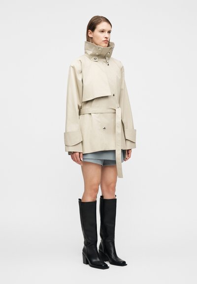 Γυναίκα που φοράει μπεζ ζακέτα με ζώνη και ψηλό γιακά, ανοιχτό μπλε σορτς και μαύρες μπότες με τακούνι που φτάνουν μέχρι το γόνατο, στέκεται μπροστά σε λευκό φόντο.