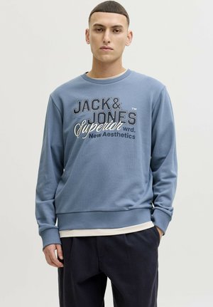 Jonge man die een blauwe trui draagt met de tekst "JACK & JONES Superior New Aesthetics", staand met één hand in de zak tegen een effen achtergrond.