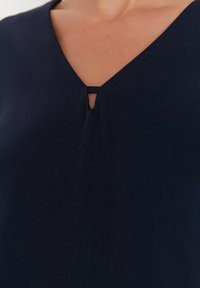 Blouse bleu marine avec un décolleté en V présentée avec un petit d�étail en forme de trou au centre. Tissu lisse avec une texture légèrement drapée.