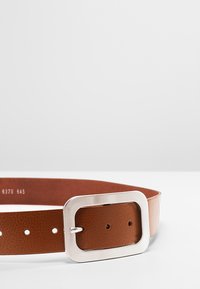 Ceinture en cuir marron avec une boucle rectangulaire en argent, présentant des trous régulièrement espacés le long de la ceinture pour ajustement. Texture lisse.