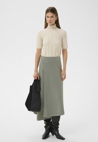 Beiges, geripptes Rollkragenshirt, grüner Wickelrock mit seitlichem Schlitz, schwarze glänzende kniehohe Stiefel und eine große schwarze Tasche.