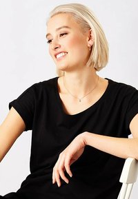 T-shirt nera con collo rotondo e maniche corte. Il modello ha capelli biondi chiari e corti, indossa una collana delicata ed è seduto in modo casual.