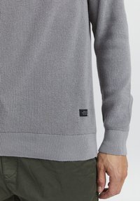 Grauer Strickpullover mit strukturiertem Muster, gerippten Bündchen und Saum. Ein kleines schwarzes Etikett ist auf der unteren linken Seite sichtbar.