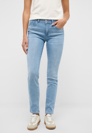 Frau trägt hellblaue eng anliegende Jeans, weiße Sneakers und ein weißes ärmelloses Oberteil, steht mit einer Hand in der Tasche.