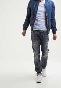 Sininen bomber-takki valkoisen t-paidan päällä, yhdistetty kuluneisiin sinisiin farkkuihin ja valkoisiin tennareihin. Paino casual-tyylillä ja rentoutuneella istuvuudella.