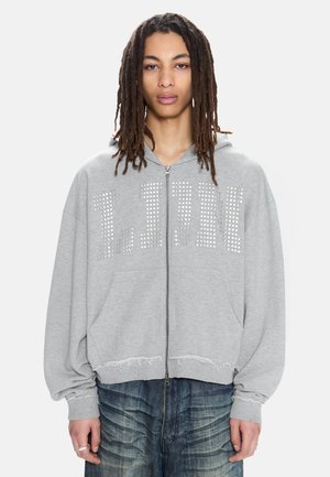 Persona joven con largas rastas que lleva una sudadera con cremallera gris con tachuelas plateadas formando "LJUN" y bajos desgastados, combinada con vaqueros azules.