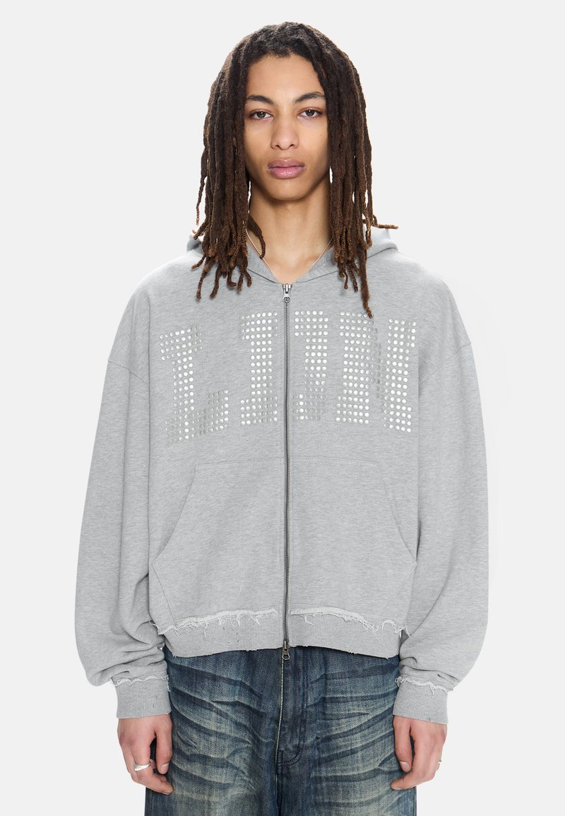 Νεαρός με μακριά dreadlocks φορά γκρι φούτερ με φερμουάρ, διακοσμημένο με ασημένια καρφιά που σχηματίζουν τη λέξη "LJUN" και φθαρμένα τελειώματα πάνω από μπλε τζιν.