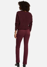 Pull en tricot bordeaux avec un col large et des boutons décoratifs sur les manches, associé à un pantalon bordeaux ajusté et des bottines noires à talons.