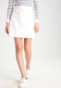 Biała spódnica w kształcie litery A, wykonana z teksturowanej tkaniny, długości powyżej kolan, zestawiona z białymi sneakersami na slip-on, noszona z pasującą, w paski bluzką z długim rękawem.