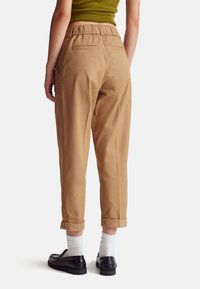 Béžové bavlněné kalhoty s volným střihem, elastickým pasem, dvěma zadními kapsami a ohrnutými konci, spárované s černými botami a bílými ponožkami.