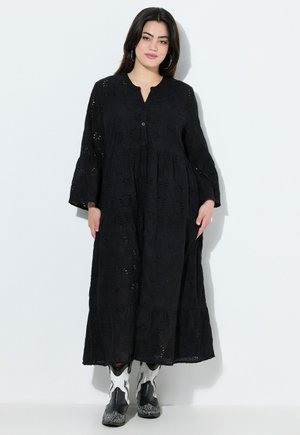 Vestido negro de manga larga con bordado de ojal, escote en V, cierre de botones, falda de capas y silueta fluida. Combinado con botas en blanco y negro.