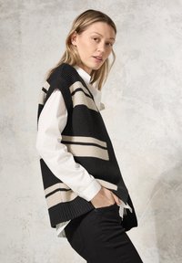 Schwarze und beigen Strickweste mit weiten Armausschnitten, die horizontale Streifen und eine strukturierte Oberfläche aufweist, getragen über einem weißen Hemd mit Kragen.