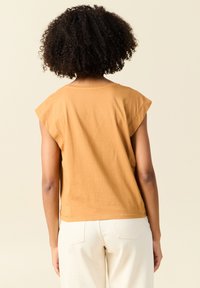 Top a maniche corte di colore senape realizzato in tessuto morbido, con scollo rotondo e vestibilità rilassata, abbinato a pantaloni chiari.