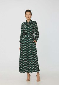 Robe longue verte avec un col et des manches longues. Présente un motif géométrique rose et blanc, une taille à nouer et des poches latérales. Tissu lisse.