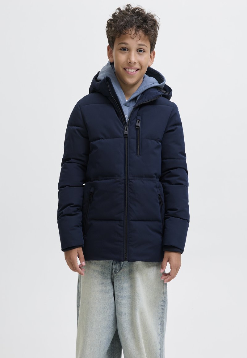 Ragazzo sorridente che indossa una giacca imbottita blu navy sopra una felpa grigia con cappuccio e jeans azzurri, in piedi di fronte a uno sfondo semplice.