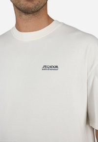 Kortærmet t-shirt med rund hals i off-white bomuld, med et blåt og sort "PEGADOR" logo og teksten "scent of summer" på venstre bryst.
