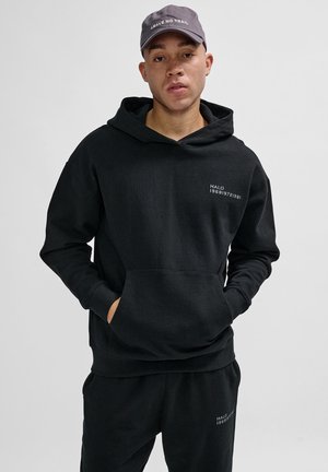 Schwarzer Hoodie mit Fronttasche, versehen mit einem kleinen weißen Logo. Kombiniert mit passenden schwarzen Hosen trägt das Modell eine graue Mütze mit Text.