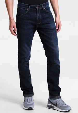 Mężczyzna ubrany w ciemnoniebieskie jeansy slim-fit i szare buty sportowe Nike stoi na tle jasnego, jednolitego tła.