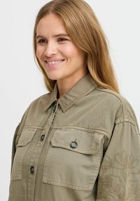 Olive zelená košová bunda z bavlnenej zmesi, s dvoma náprsními vreckami, zapínaním na gombíky a vyšitým kvetinovým detailom na rukávoch.