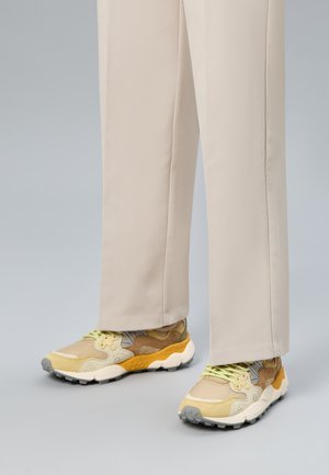 Baskets beige, jaunes et blanches avec des lacets verts, portées avec un pantalon beige clair droit sur un sol gris.