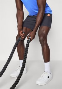 Mand i blå skjorte og sorte shorts træner med sorte battle ropes, iført hvide sokker og hvide sneakers på grå gulv.