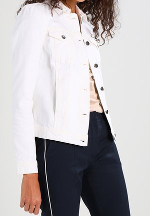 Mujer con una chaqueta blanca abotonada sobre una camiseta beige y pantalones azul marino con una raya blanca lateral, mostrada desde el hombro hasta la cintura.