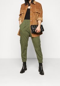 Vrouw in een bruine corduroy overshirt, zwarte top, olijfgroene cargobroek, zwarte hoge laarzen, met een zwarte gequiltte clutch in haar hand.
