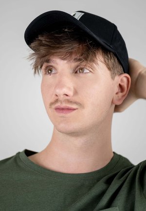 Black cap with a green shirt, featuring a round neckline. Short, tousled hair and a subtle mustache. Neutral background.