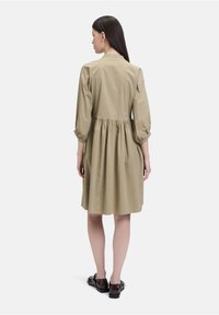 Khakifarbens Baumwollkleid mit tailliertem Oberteil und lockerem, gerafftem Rock; mit Puffärmeln und rundem Ausschnitt; kombiniert mit glänzenden braunen Schuhen.