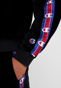 Veste en corduroy noir avec un design rayé rouge et bleu le long des manches, rehaussée de logos blancs. Poignets côtelés pour un ajustement parfait.