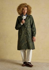 Žalias vandeniui atsparus paltas su gėlių raštu, užtrauktuku priekyje, reguliuojama kepure ir dviem priekinėmis kišenėmis. Su derančiomis baltomis kelnėmis ir rudos spalvos sportbačiais.