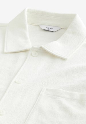 Camicia bianca con colletto, in tessuto strutturato e vestibilità rilassata, caratterizzata da bottoni sul davanti e una tasca sul lato sinistro.