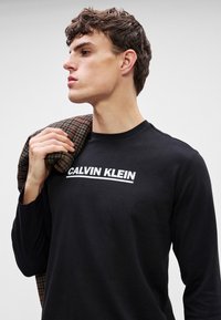 Camisa de mangas longas preta com o logótipo branco "CALVIN KLEIN". Material de algodão suave, design ajustado, usada com um casaco xadrez castanho sobre o ombro.