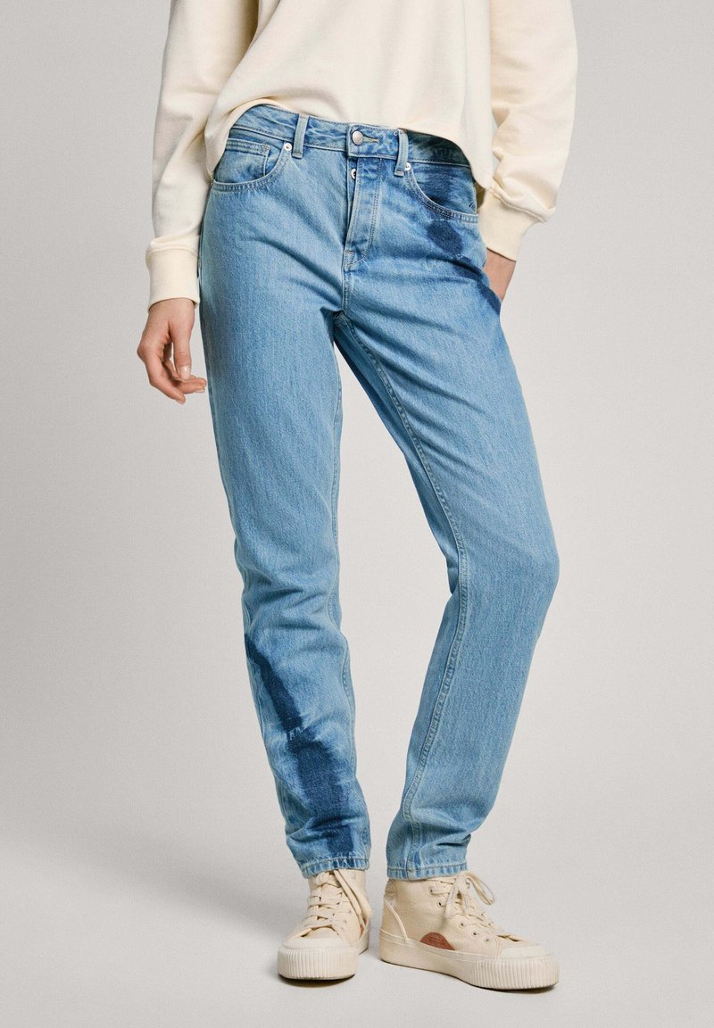 Jeans de denim azul claro com um corte descontraído, apresentando um acabamento desbotado e uma mancha azul escura subtil na perna esquerda, combinados com ténis bege.