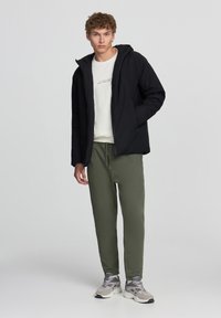Giacca a vento nera con cappuccio, indossata sopra un maglione crema, abbinata a pantaloni sportivi verde oliva e sneakers grigie.