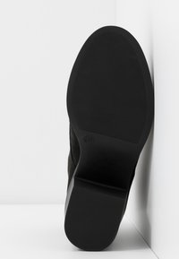 Bottine en daim noir avec une semelle en caoutchouc texturée et un bout arrondi. Elle présente un talon plat et un design simple, sans ornements.