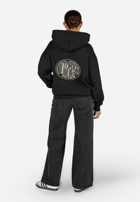 Juodas džemperis su dideliu apvaliu grafikiniu piešiniu nugarinėje pusėje, kuriame pirminis tekstas yra baltos ir aukso spalvų. Laidūs kirpimai, erdvus kapišonas ir riboti rankogaliai. Derinama su plačiomis pilkomis džinsomis.