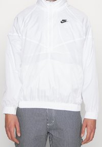 Vit lättviktsjacka med blixtlås och meshstrukturer, med hög krage och elastiska manschetter. Svart Nike-logga på vänster bröst. Randiga byxor synliga.