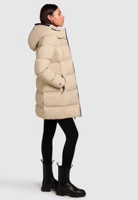 Bézs pufi kabát kapucnival, elülső cipzárral és fekete részletekkel, fekete leggingsszel és térdig érő fekete csizmával párosítva.