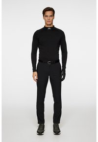 Svart långärmad tröja med hög krage och logotyp, tillsammans med svarta tighta byxor. Modellen bär svarta handskar och svarta sneakers för en komplett outfit.