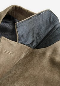 Tissu en daim marron avec une texture lisse, doté d'un col rabattu. L'intérieur présente une doublure à motifs gris foncé et des accents en tissu noir uni.