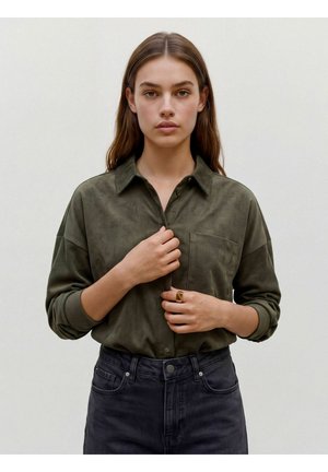 Jeune femme aux longs cheveux bruns portant une chemise en velours côtelé verte et un pantalon noir, boutonnant sa chemise avec une bague en or à son doigt.
