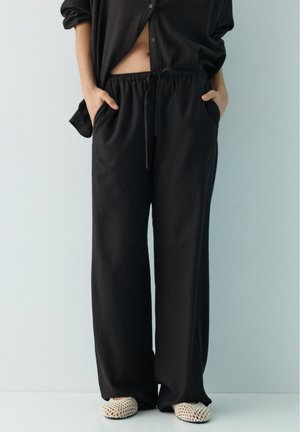 Person iført løse sorte bukser med snøre og sort skjorte, hænder i lommerne, stående mod en lysegrå baggrund.