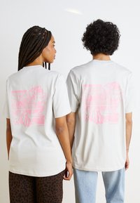 Ljust grå bomullströjor med korta ärmar, med en stor rosa grafisk design på baksidan. Tyget verkar mjukt med en avslappnad passform.