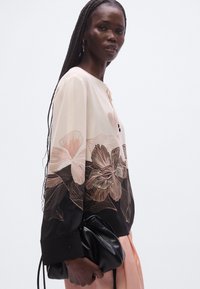 Blouse à manches longues avec un corps crème orné de motifs floraux roses et noirs, manches noires contrastantes et détails de boutons aux poignets.