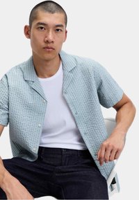 Jongeman met kort haar in een lichtblauw, gestructureerd, kort mouw shirt over een witte tanktop, zittend op een witte stoel.