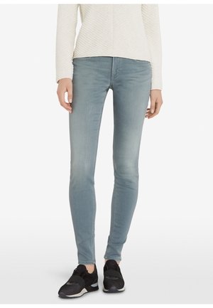 Jean skinny bleu clair en denim, avec une texture lisse et un design ajusté. Porté avec un haut blanc texturé et des baskets noires.