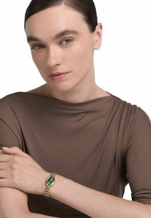 Frau mit zurückgekämmtem Haar trägt ein braunes Oberteil und eine goldene Armbanduhr mit grünem Zifferblatt und posiert mit verschränkten Armen vor weißem Hintergrund.